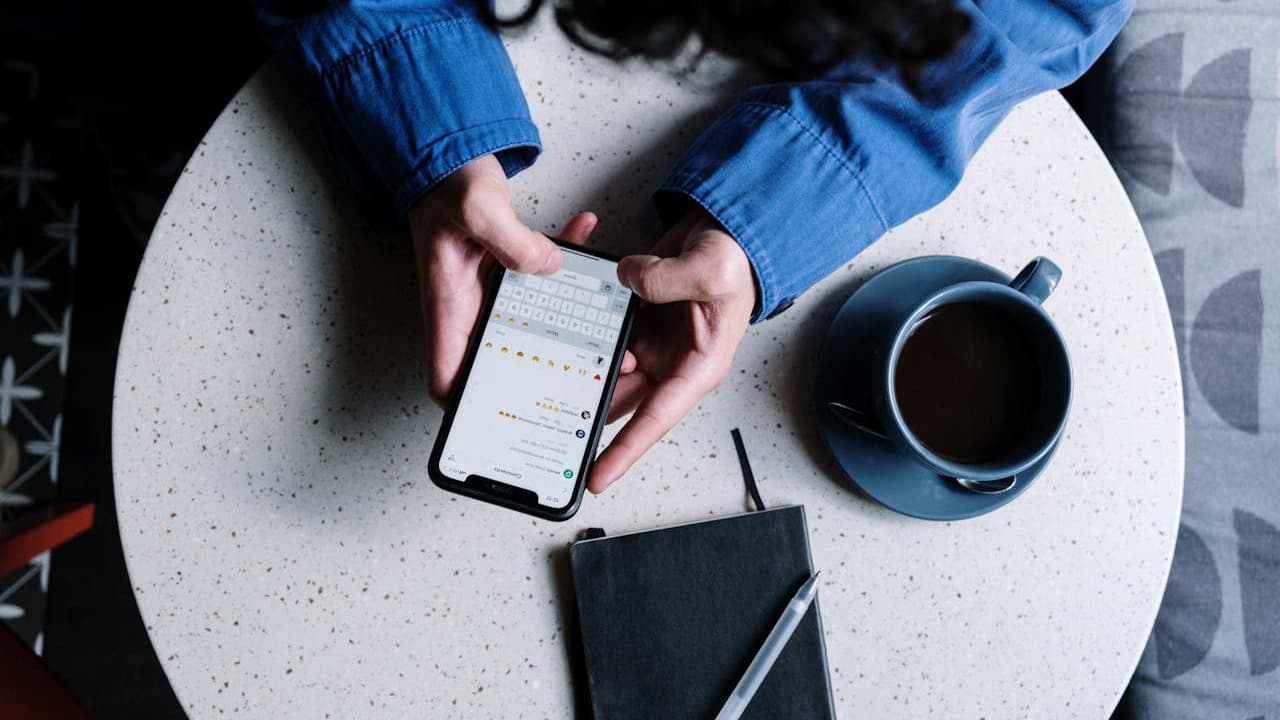 Person typing on a smartphone at a round table, blue coffee cup, black notebook, and pen beside them