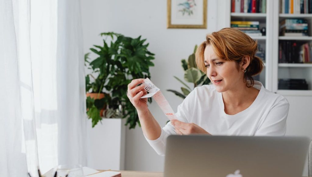 Woman holding a long receipt, looking concerned, sitting at a desk with a laptop in a home office setting