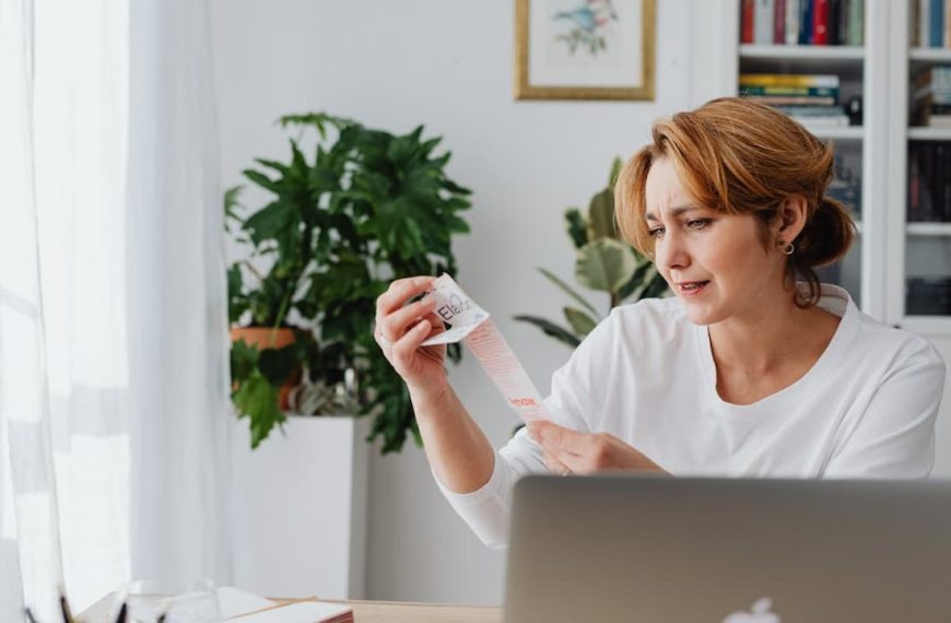 Woman holding a long receipt, looking concerned, sitting at a desk with a laptop in a home office setting