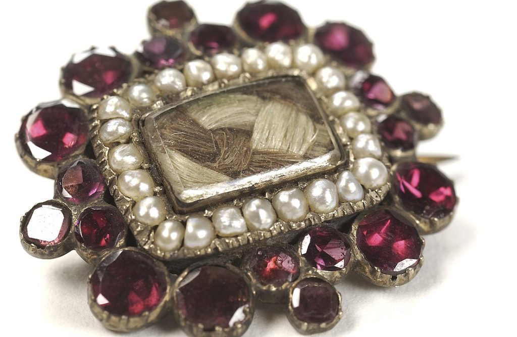 Vintage brooch with a large rectangular central gemstone, surrounded by a row of white pearls, encircled by deep red round gemstones, set in an ornate gold-toned metal