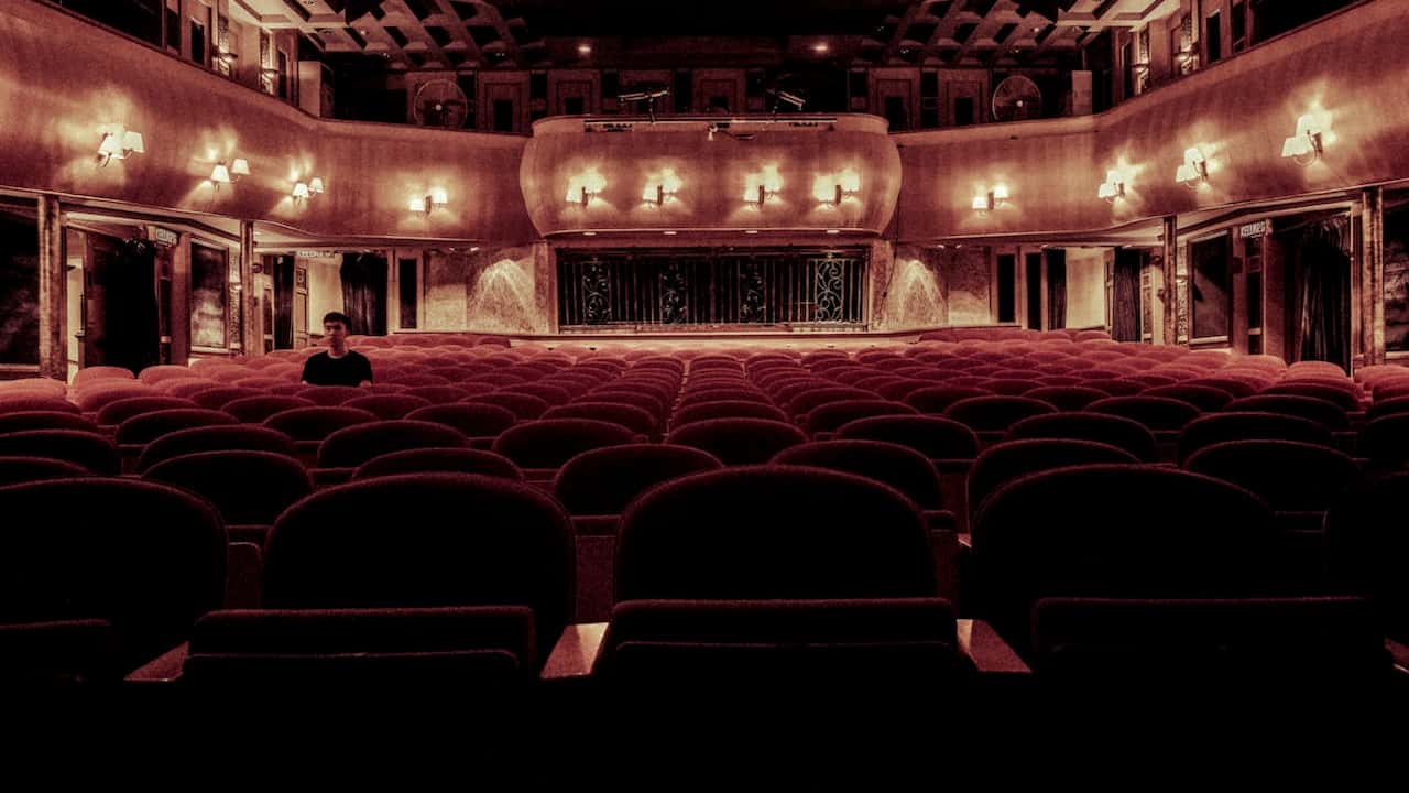Dimly lit vintage-style theater with rows of red velvet seats, ornate lighting, and a single person seated near the front, facing the empty stage