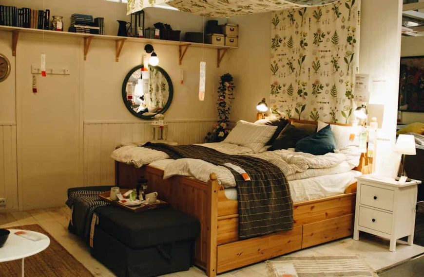 A cozy bedroom display with a wooden bed frame, striped bedding, and multiple light sources including bedside lamps, wall sconces, and a ceiling pendant. The space is decorated with botanical-patterned curtains, a round mirror, and a floating shelf with books and deco