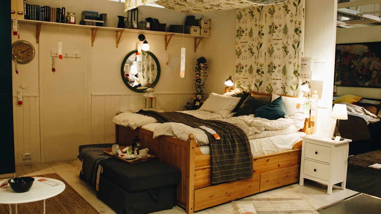 A cozy bedroom display with a wooden bed frame, striped bedding, and multiple light sources including bedside lamps, wall sconces, and a ceiling pendant. The space is decorated with botanical-patterned curtains, a round mirror, and a floating shelf with books and deco