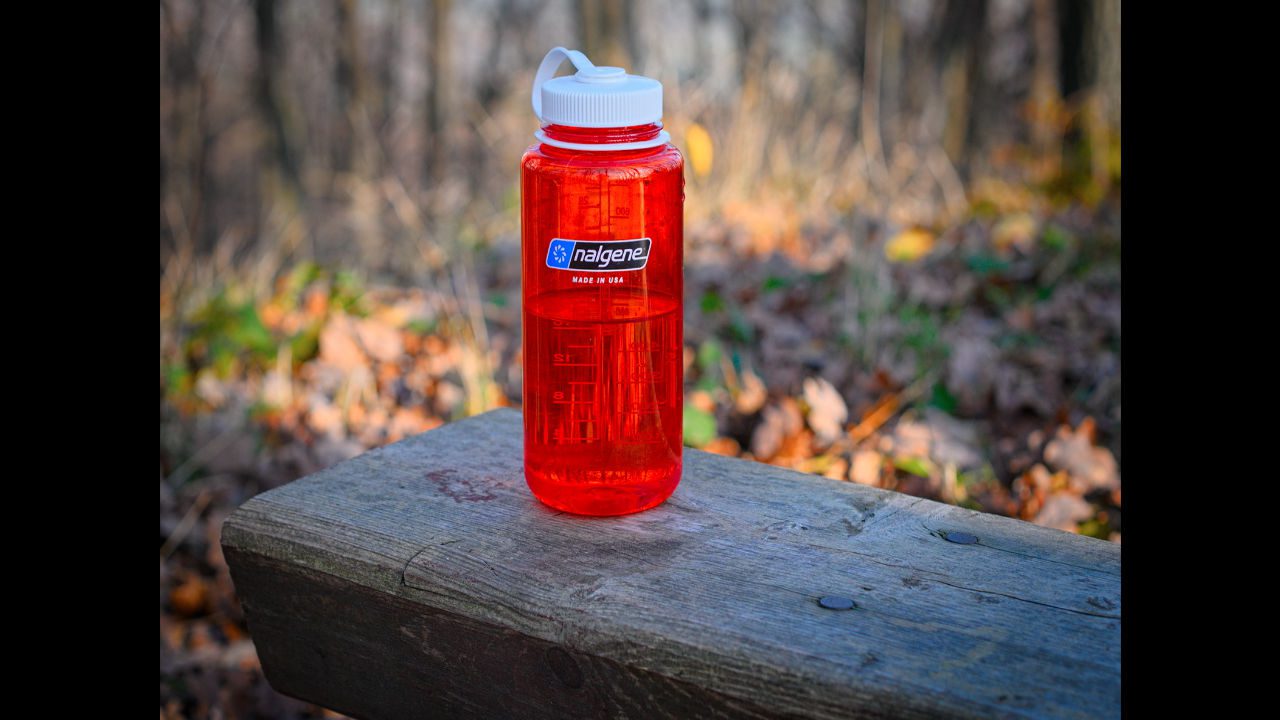 Red Nalgene wide-mouth water bottle with white lid placed on a wooden bench in an outdoor setting