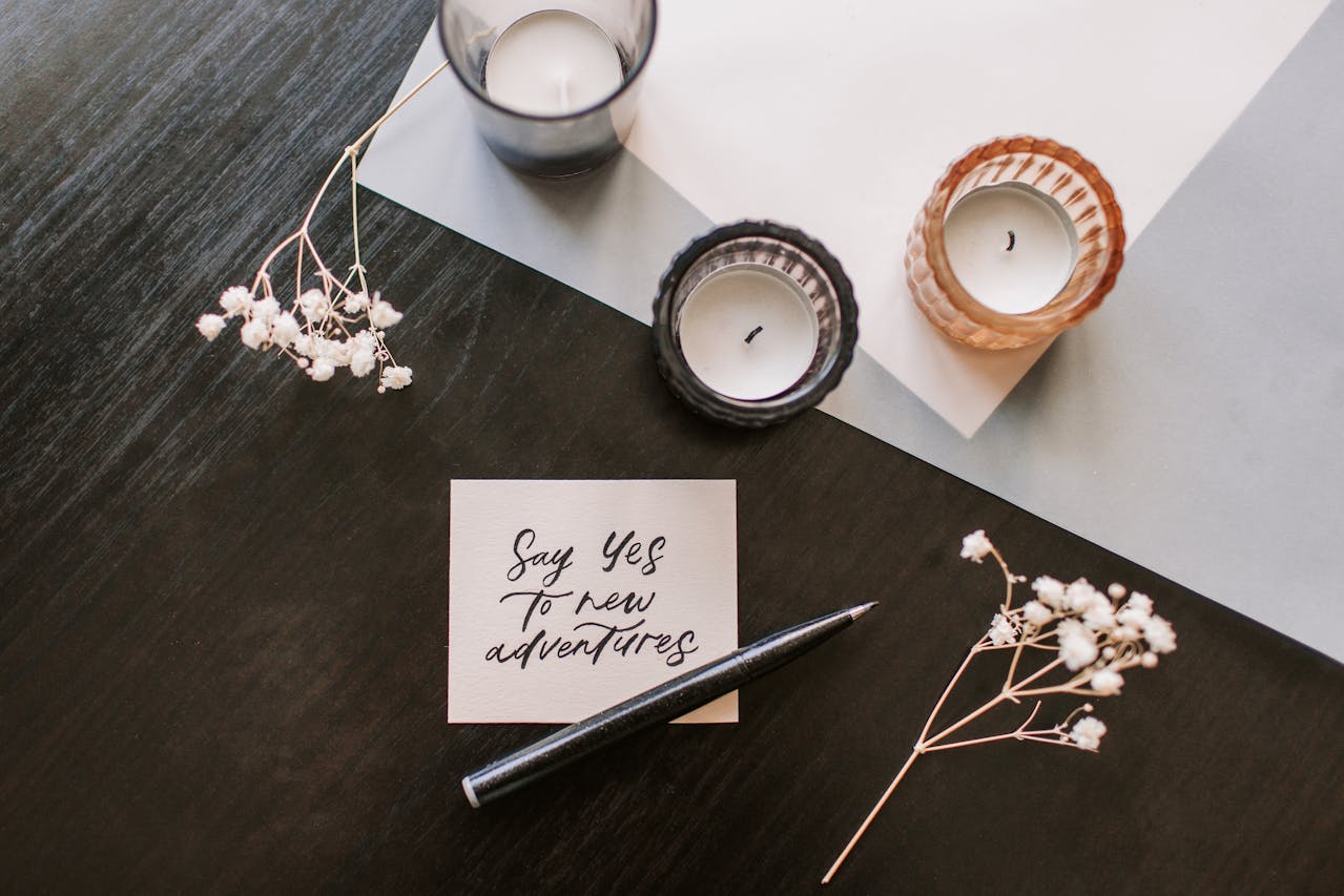 Handwritten note reading "Say yes to new adventures", black pen beside the note, three lit candles in glass holders, sprigs of white dried flowers, dark wooden table, angled white paper beneath items