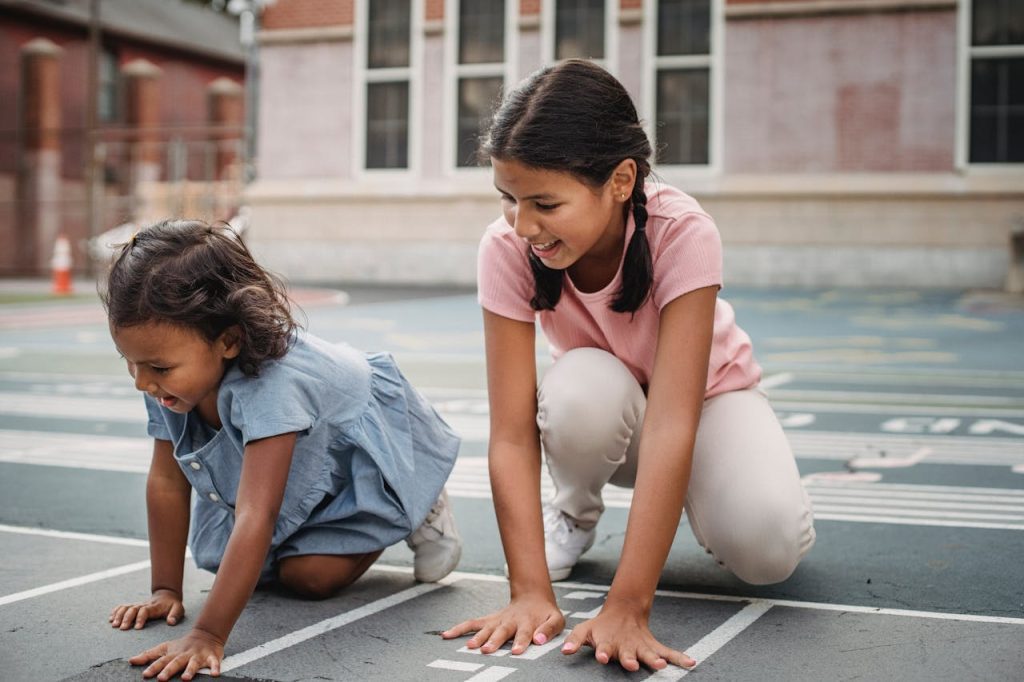 two young girls playing and laughing on a painted outdoor track in a schoolyard or neighborhood playground