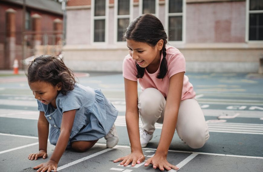 two young girls playing and laughing on a painted outdoor track in a schoolyard or neighborhood playground