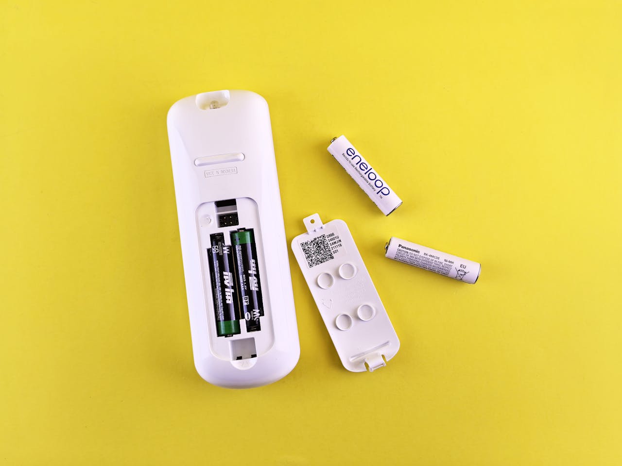 White remote control with its battery cover removed, two AA batteries lying next to it, placed on a bright yellow background, exposing the battery compartment and internal components