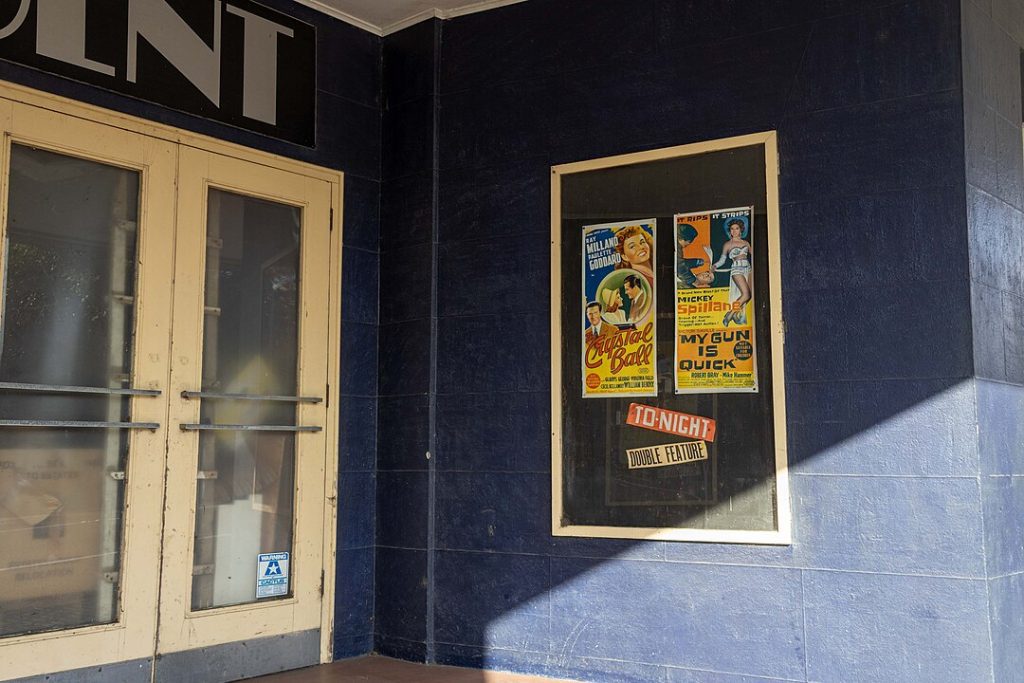 Double glass doors with reflections, dark blue wall, framed movie posters displayed in a glass case, partial shadow cast across the wall
