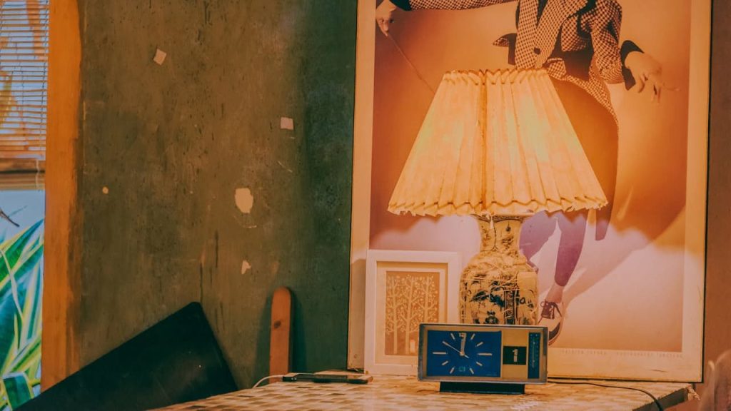 A vintage ceramic table lamp with a pleated beige lampshade sits on a mosaic-tiled table, accompanied by a retro blue clock and framed artwork, against a textured green wall and a large fashion poster backdrop