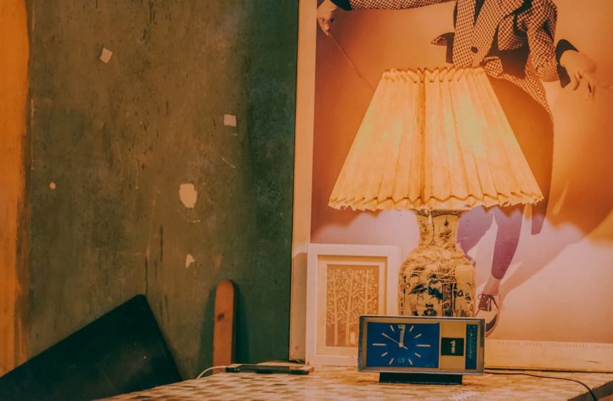 A vintage ceramic table lamp with a pleated beige lampshade sits on a mosaic-tiled table, accompanied by a retro blue clock and framed artwork, against a textured green wall and a large fashion poster backdrop