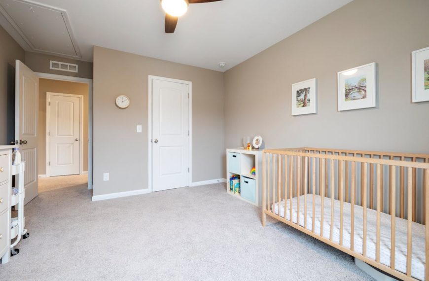 Minimalist room, light beige walls, wooden crib with no bedding, soft carpeted floor, small white toy storage unit, three framed pictures on the wall, ceiling fan with light
