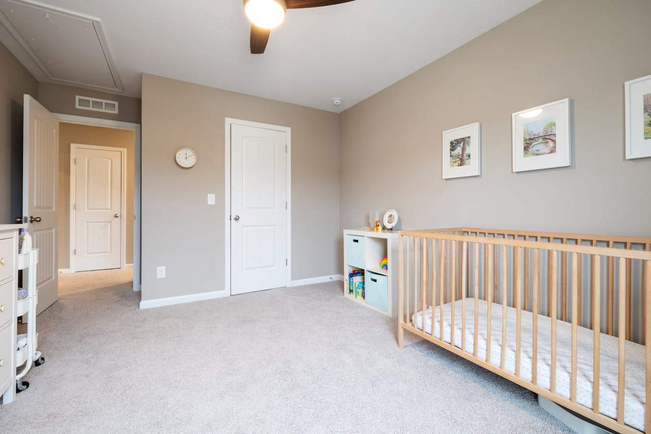 Minimalist room, light beige walls, wooden crib with no bedding, soft carpeted floor, small white toy storage unit, three framed pictures on the wall, ceiling fan with light