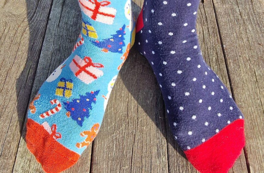 Mismatched socks on feet, left sock is blue with Christmas trees and gifts, right sock is dark blue with white polka dots, both socks have colorful toes, worn on wooden floor