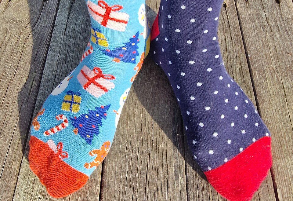 Mismatched socks on feet, left sock is blue with Christmas trees and gifts, right sock is dark blue with white polka dots, both socks have colorful toes, worn on wooden floor