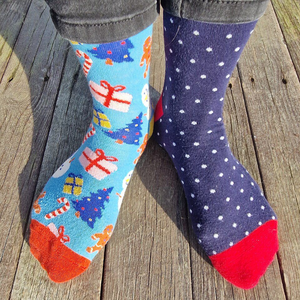 Mismatched socks on feet, left sock is blue with Christmas trees and gifts, right sock is dark blue with white polka dots, both socks have colorful toes, worn on wooden floor