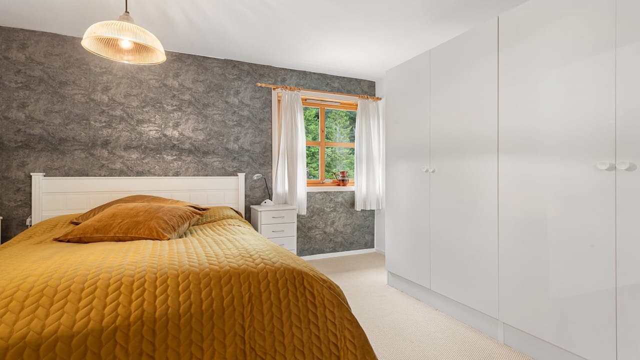 Bedroom with oversized white wardrobe, bed with golden quilt and brown pillows, textured dark gray accent wall, window with white curtains, and a white nightstand with a lamp