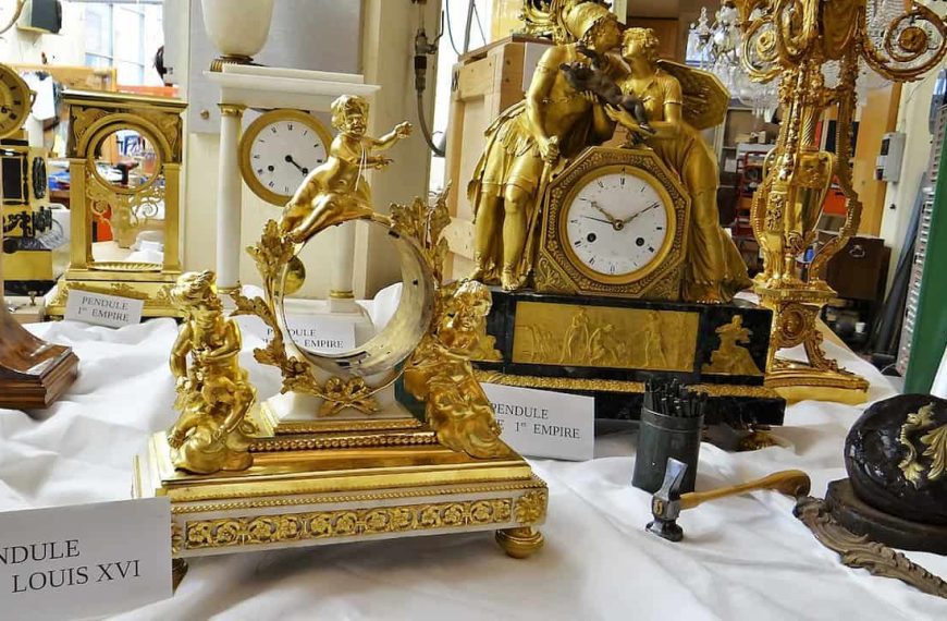Collection of ornate French figural bronze clocks on display, featuring gilded sculptures of cherubs, classical figures, and intricate decorative details, with signs indicating periods such as "Pendule Époque Louis XVI" and "Pendule 1er Empire