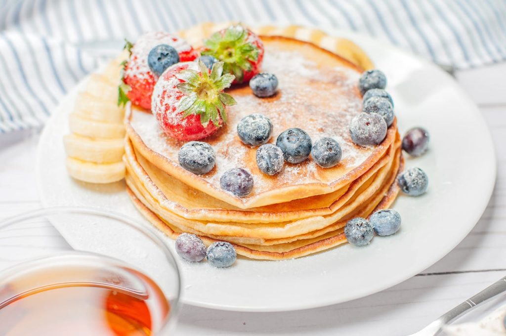 A stack of golden pancakes topped with powdered sugar, fresh blueberries, and strawberries sits on a white plate, with banana slices on the side. The setting includes a striped cloth and a glass of syrup in the foreground