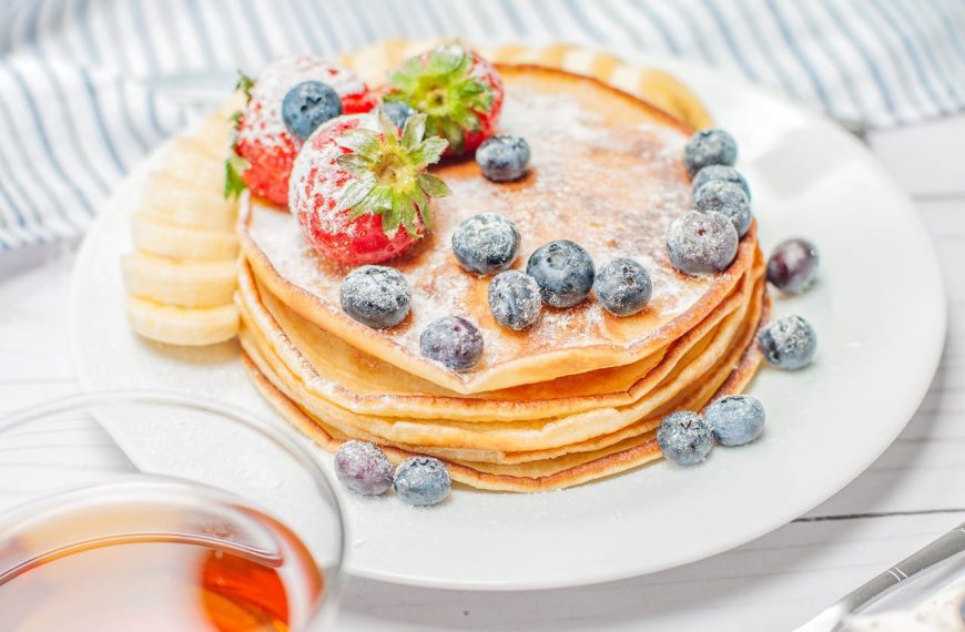 A stack of golden pancakes topped with powdered sugar, fresh blueberries, and strawberries sits on a white plate, with banana slices on the side. The setting includes a striped cloth and a glass of syrup in the foreground