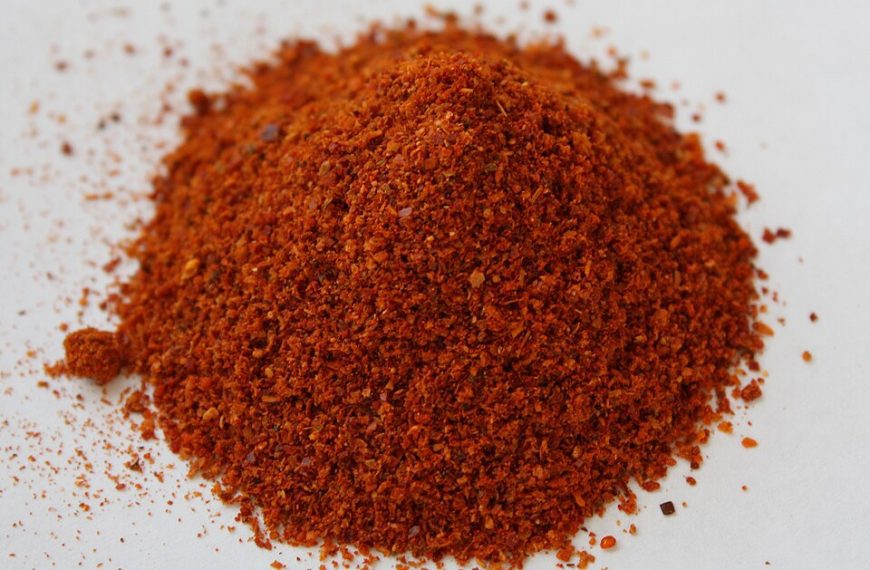 Ground red chili powder on a white surface, finely milled texture, vibrant red-orange color, commonly used spice in cooking, strong aroma, adds heat and flavor to dishes