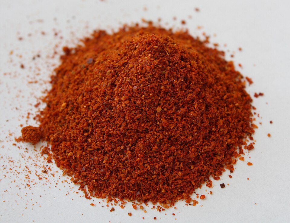 Ground red chili powder on a white surface, finely milled texture, vibrant red-orange color, commonly used spice in cooking, strong aroma, adds heat and flavor to dishes