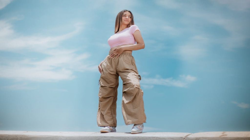Woman standing outdoors under blue sky, wearing tan parachute pants, pink fitted crop top, and white sneakers, hands resting on waist, looking confidently into the distance