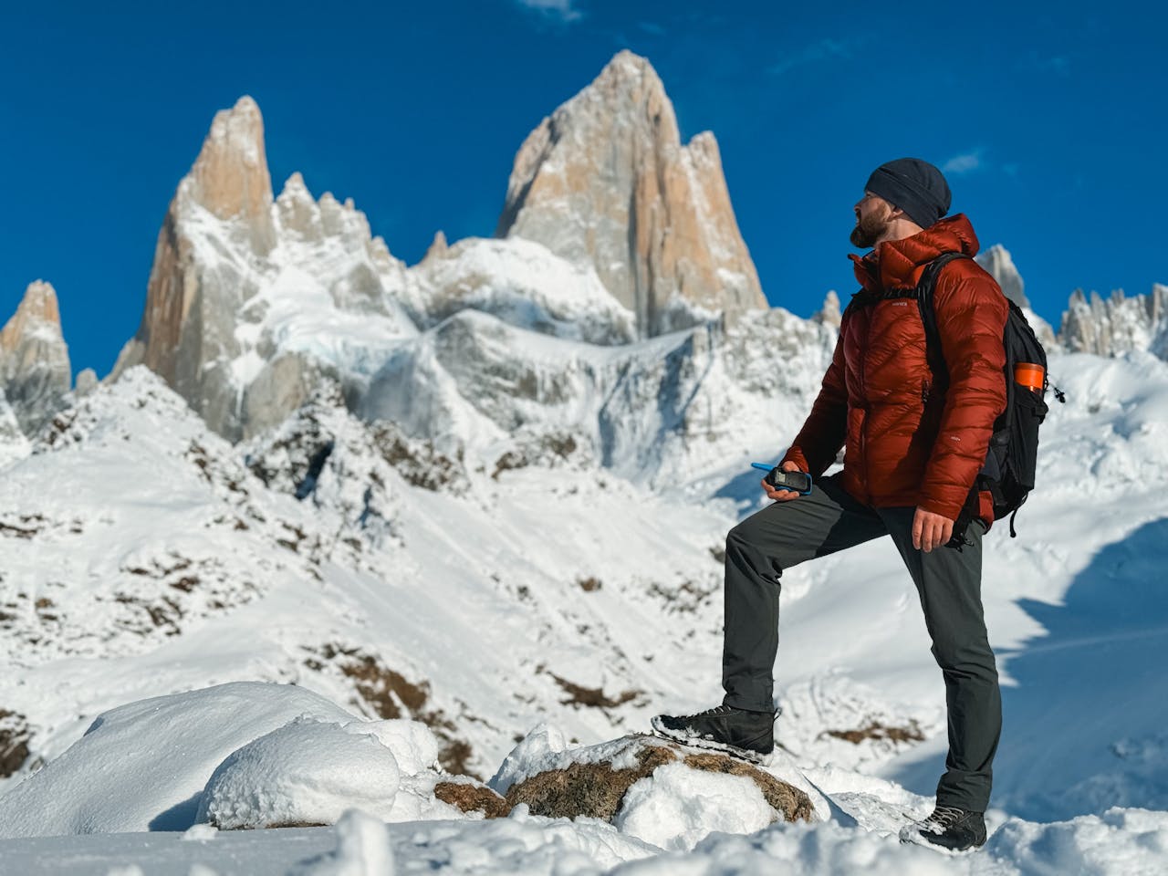 Man in a red insulated jacket and black backpack standing on a snow-covered mountain, holding a smartphone, with towering snow-covered peaks and a clear blue sky in the background