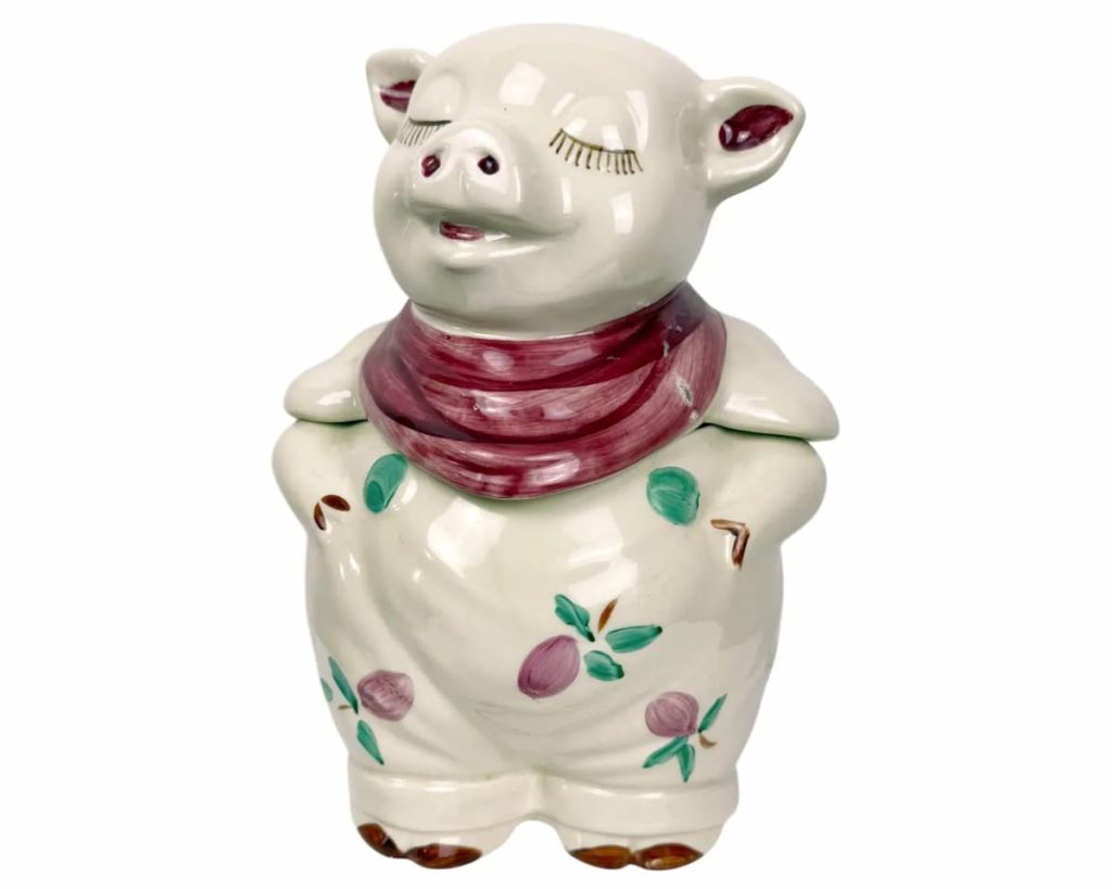 Decorative ceramic pig cookie jar with white glazed body, painted red scarf, closed eyes, and purple fruit with green leaves pattern. Standing upright with a smiling expression
