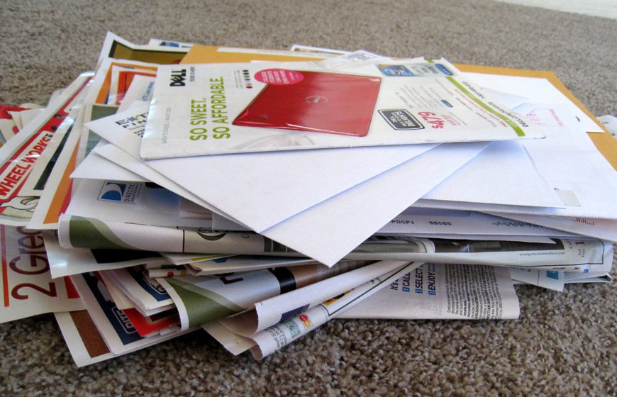 Pile of mixed mail and advertisements scattered on a carpet, including envelopes, flyers, and colorful brochures