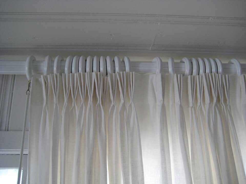Close-up of white pleated curtains, hung on a concealed curtain rod, gathered in uniform folds, mounted beneath a white ceiling cornice, softly lit by natural light
