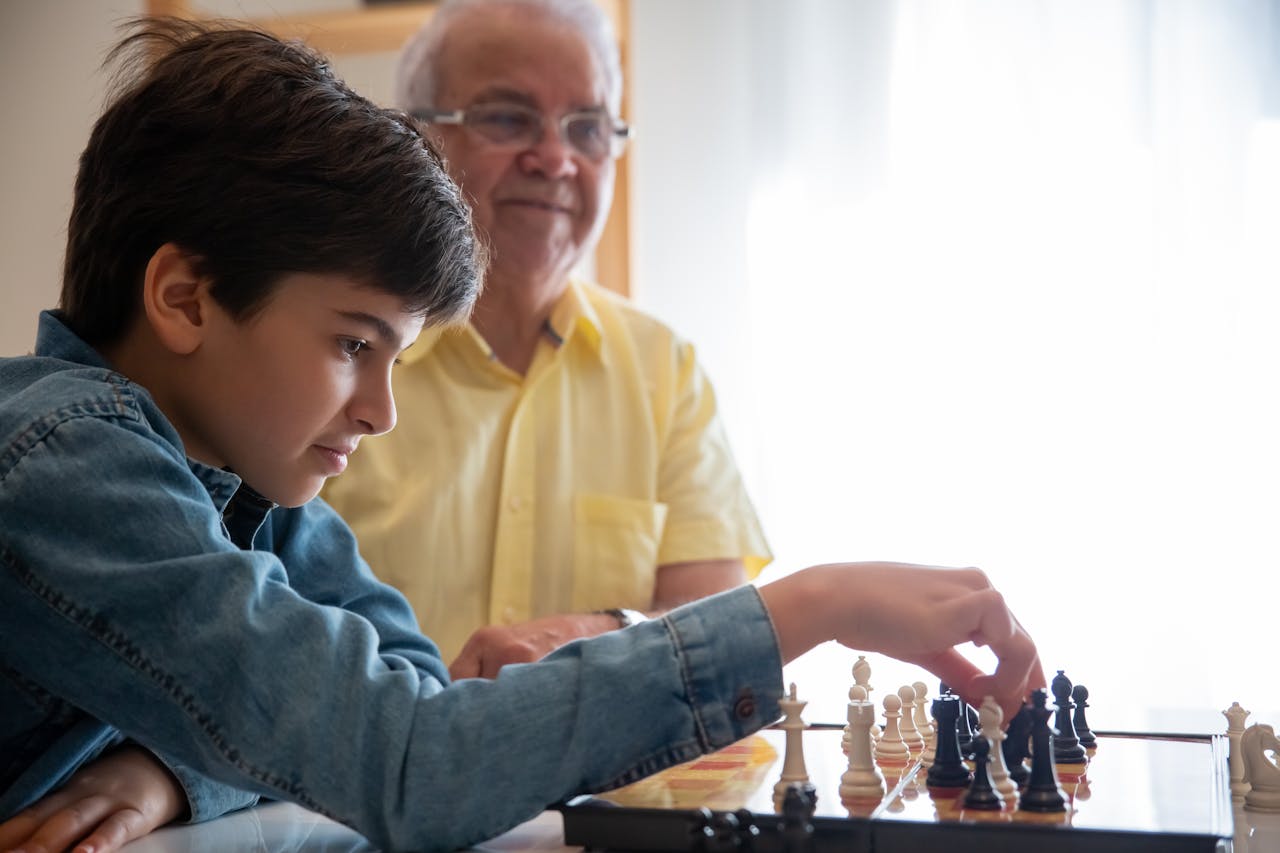 young boy in denim shirt playing chess with an elderly man in yellow shirt, focused on moving a black chess piece