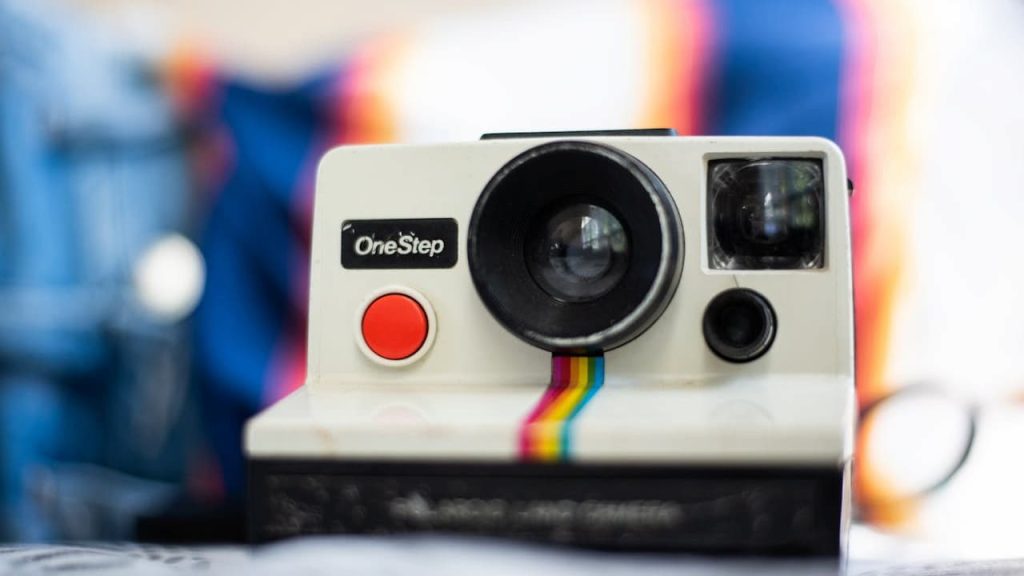Vintage Polaroid OneStep instant camera with a red shutter button and rainbow stripe, set against a colorful blurred background