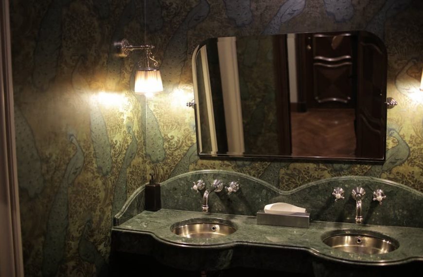 Dimly lit bathroom with a green marble double sink, ornate wall lamps, a decorative mirror, and peacock-patterned wallpaper