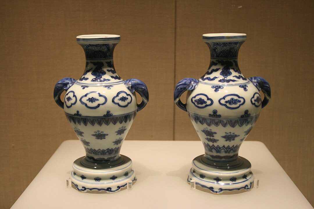 Two intricately designed blue and white ceramic vases, symmetrical floral patterns, dual handles, displayed on a beige pedestal, traditional craftsmanship.