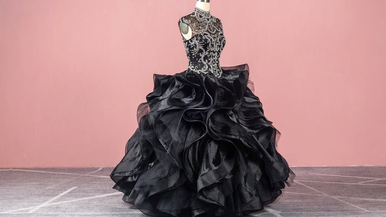 Black ball gown with a beaded bodice, high neckline, sleeveless cut, and voluminous ruffled skirt, displayed on a mannequin, pink background