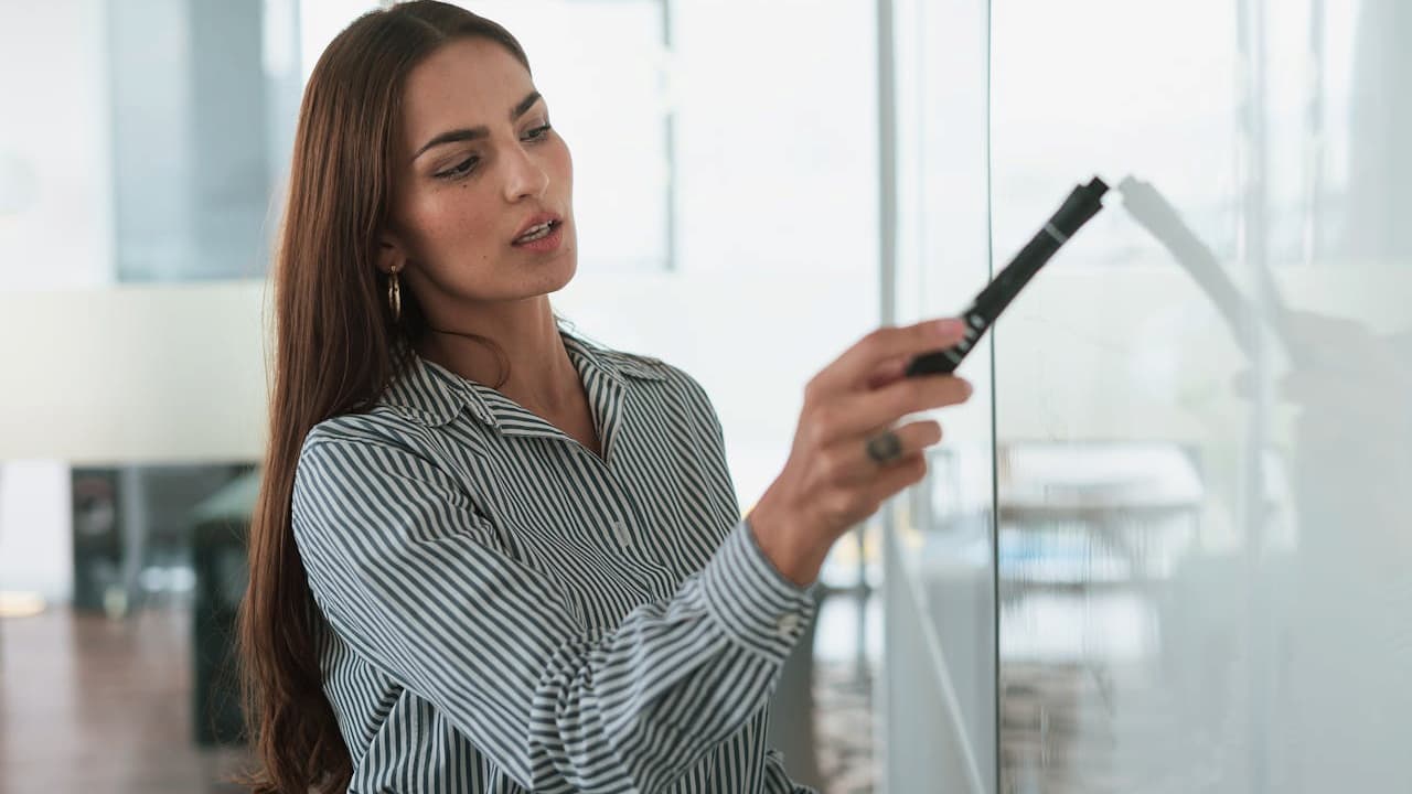 Woman in a striped button-down shirt, holding a black marker, writing on a glass whiteboard in a modern office setting
