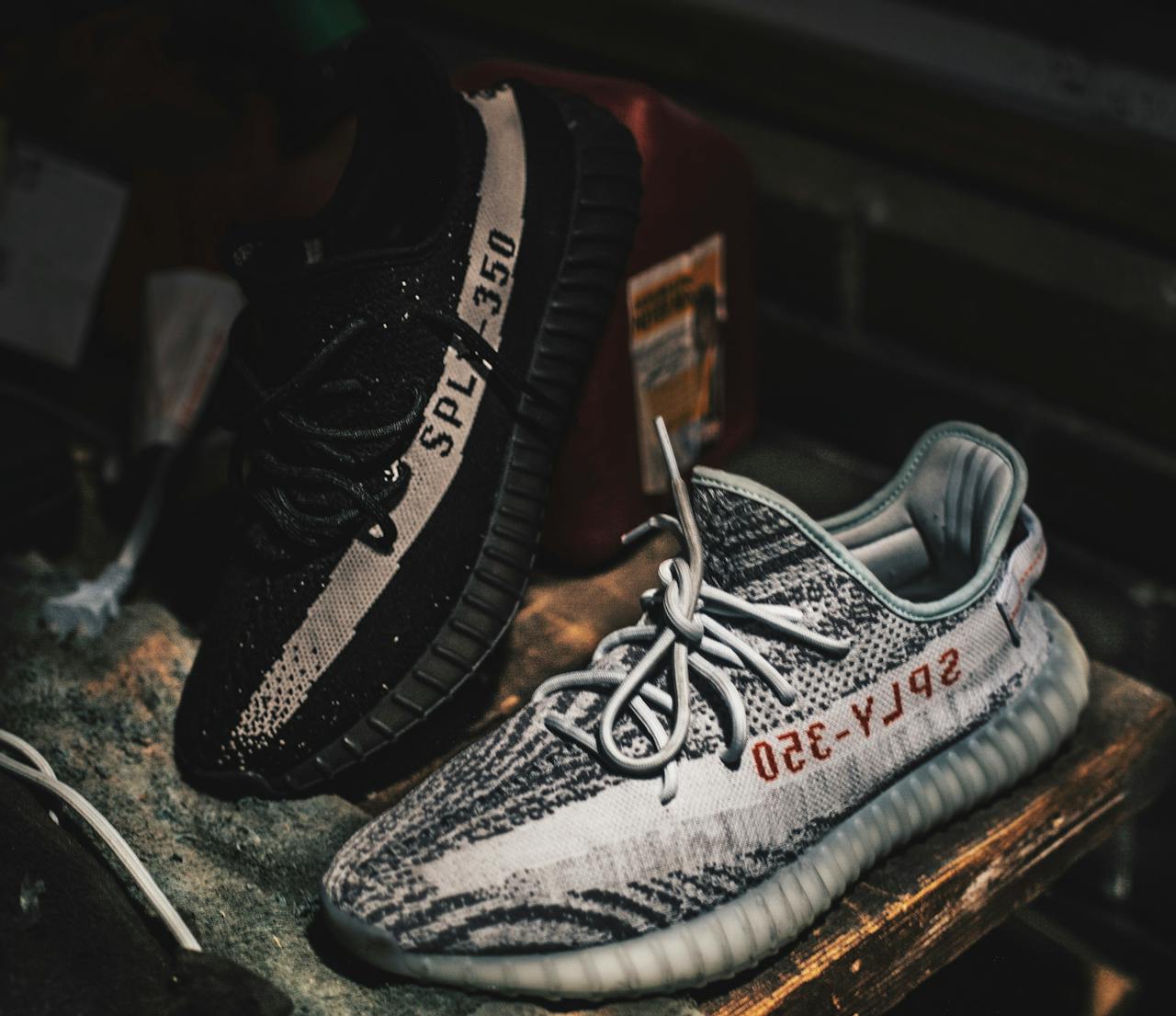 Pair of Yeezy Boost 350 V2 sneakers, one black with cream stripe, one grey with red text, displayed on rustic surface, low-light setting