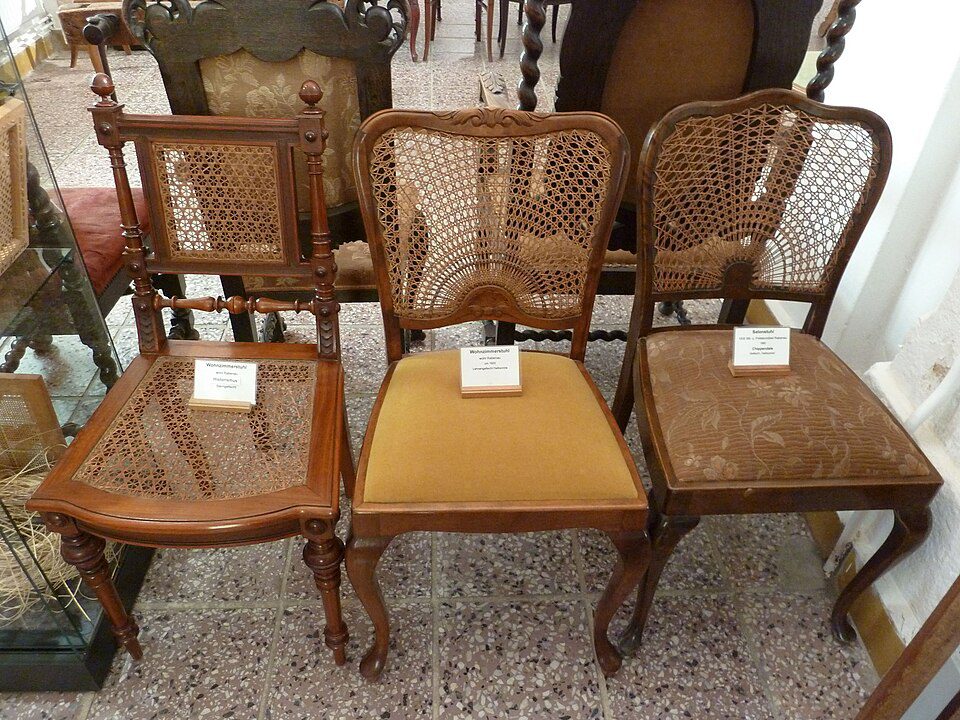 Three antique wooden chairs, each with distinct woven cane backrests, varying seat styles including wood, upholstered, and fabric, placed on a tiled floor indoors, labeled for display or sale, vintage furniture setting