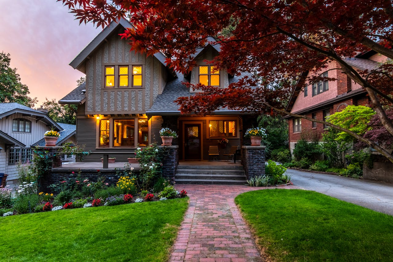 Charming craftsman-style house with glowing windows, colorful garden, red brick walkway, and vibrant surrounding plants at dusk