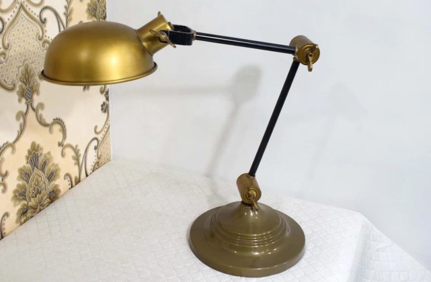 A vintage-style desk lamp with a brass dome shade and base, featuring an adjustable black metal arm with brass joints, placed on a white textured tablecloth near a wall with ornate, floral-patterned wallpaper