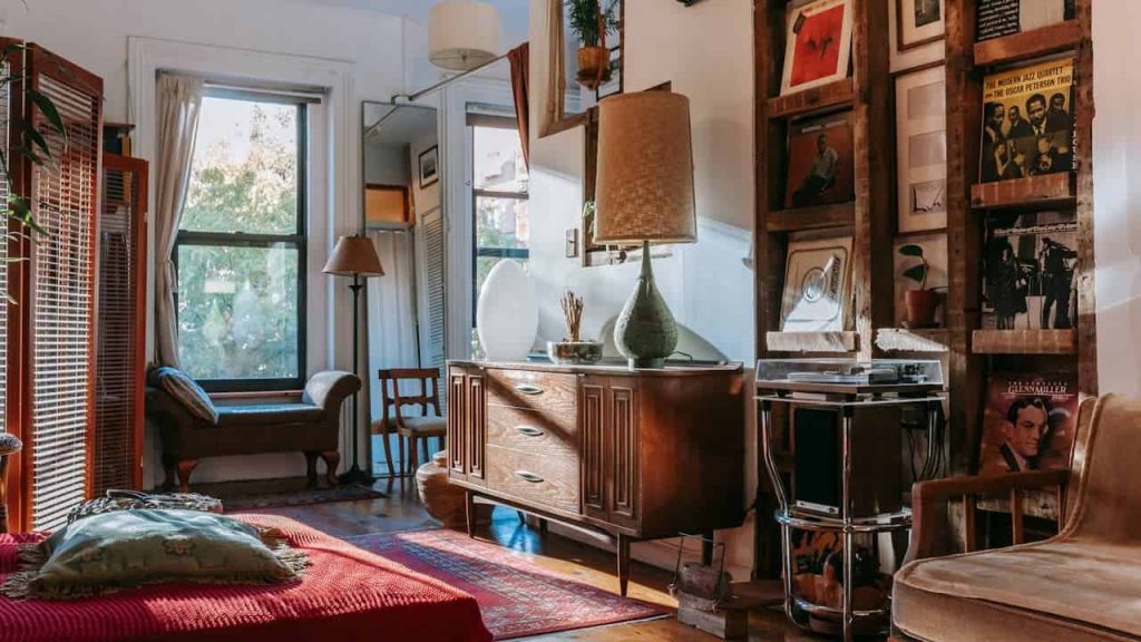 Retro-themed living room, vintage wooden sideboard, record player, vinyl records on display, red blanket-covered daybed, large windows with cream curtains, wooden floor, eclectic decor