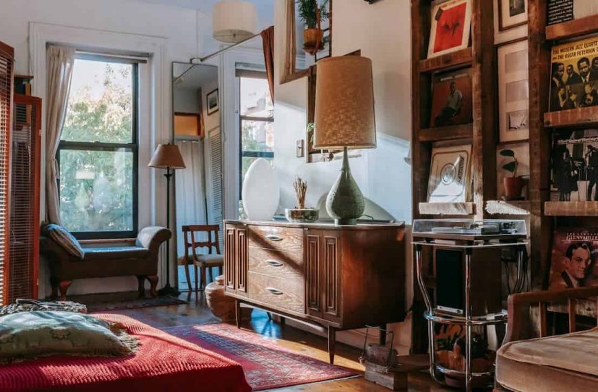 Retro-themed living room, vintage wooden sideboard, record player, vinyl records on display, red blanket-covered daybed, large windows with cream curtains, wooden floor, eclectic decor