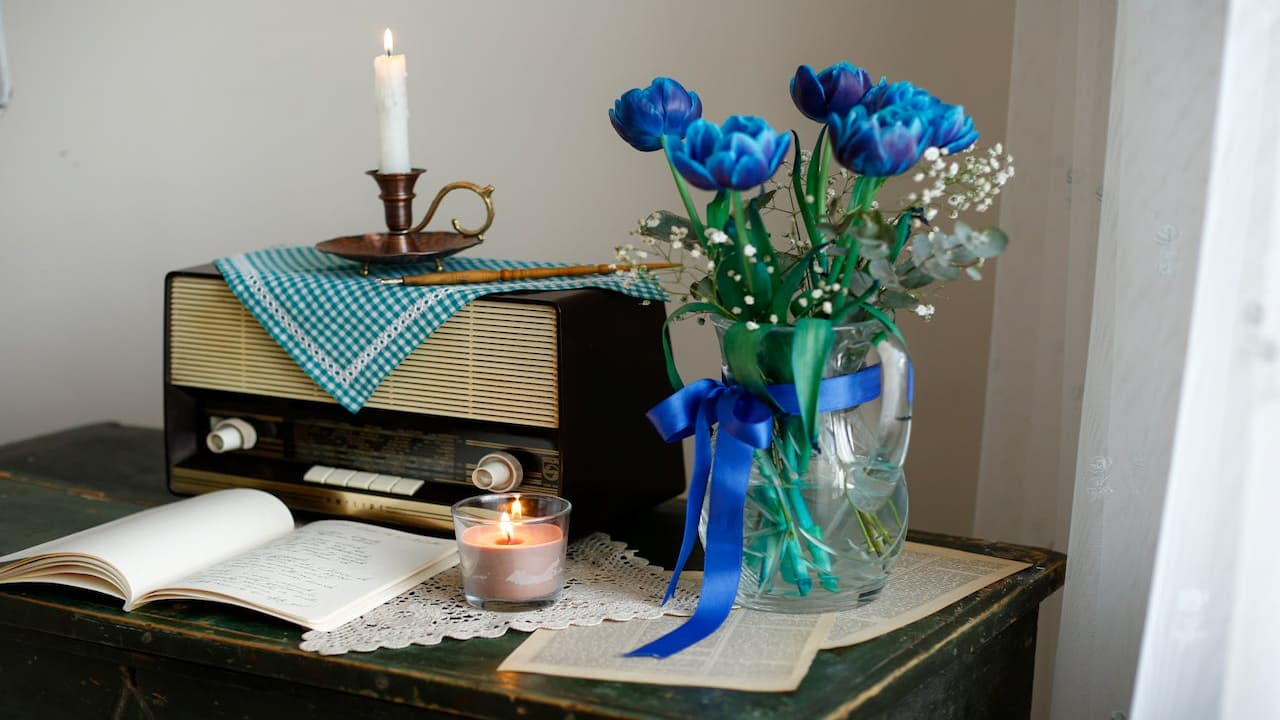 Cozy tabletop scene, blue roses in a glass vase with a blue ribbon, vintage radio with a teal cloth, open book, lit candle in a jar, candleholder with a white candle