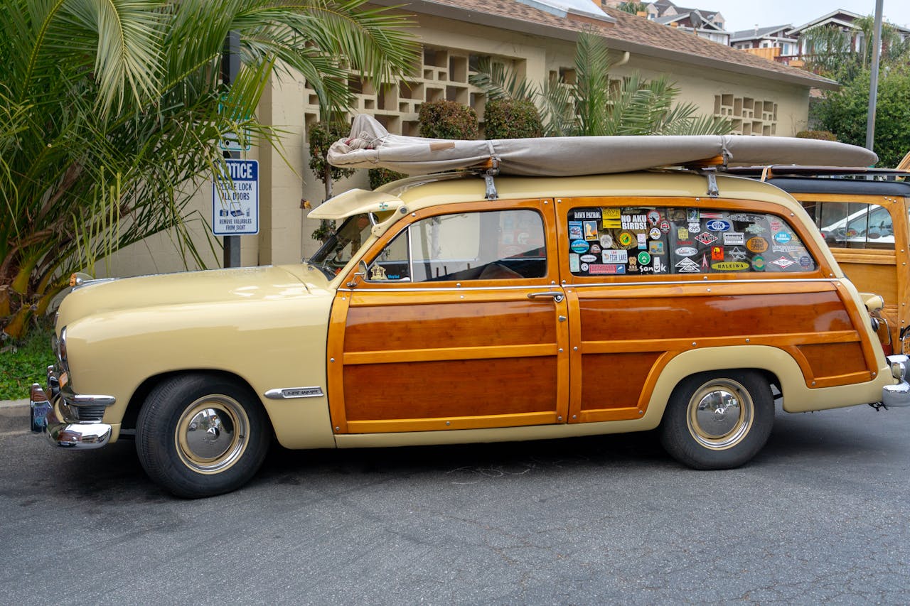 vintage station wagon with wood paneling and roof-mounted surfboard, side windows covered in travel stickers, parked near palm trees
