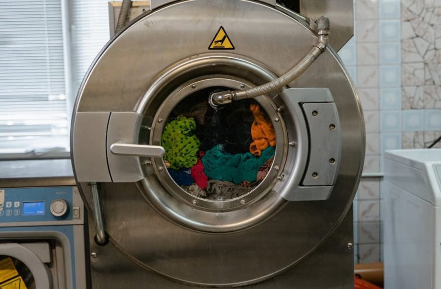 A stainless steel industrial washing machine filled with colorful clothes, viewed through the round glass door during the rinse and spin cycle in a laundry facility