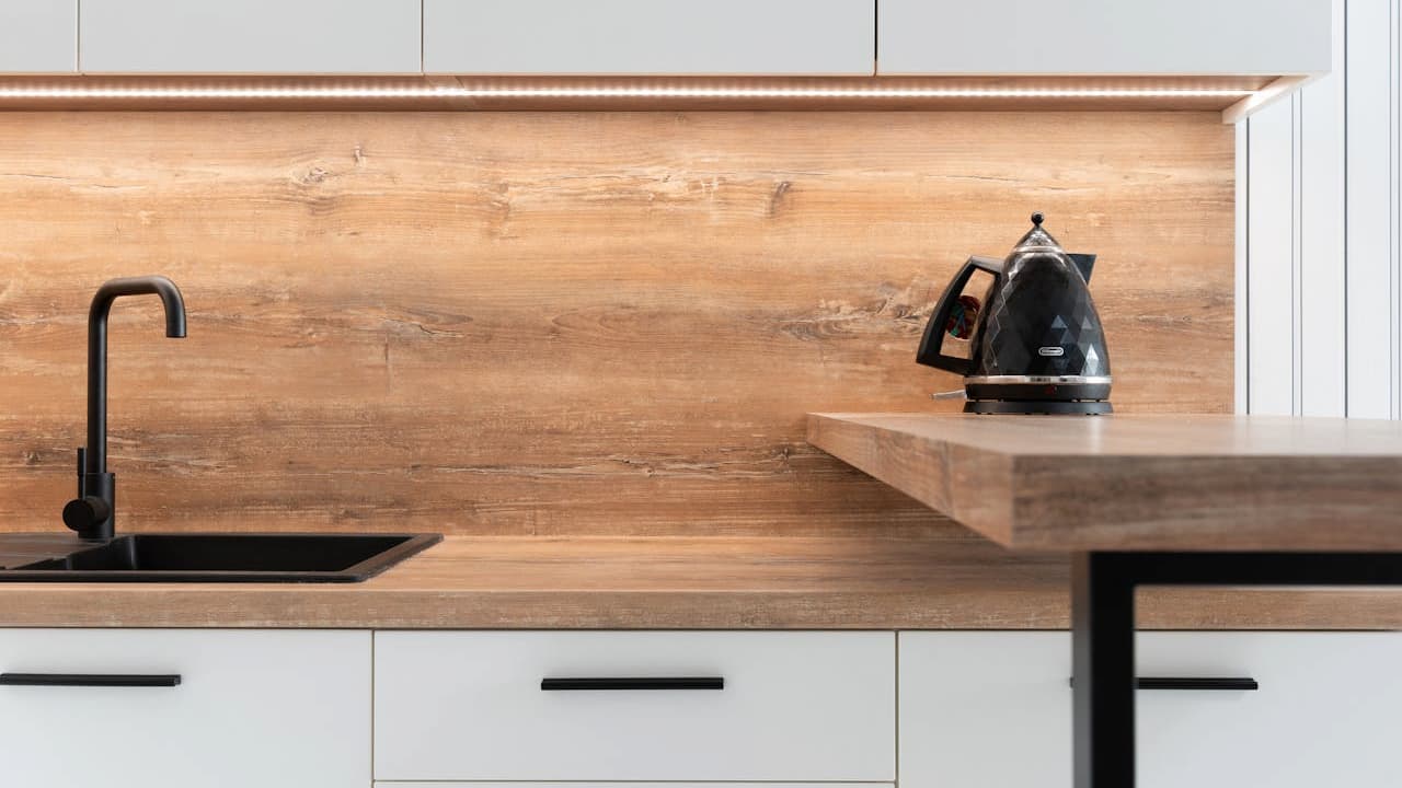 Black electric kettle on a wooden countertop, modern kitchen setup, light wood backsplash, black faucet and sink, white cabinets with black handles, minimalistic design, clean and organized space