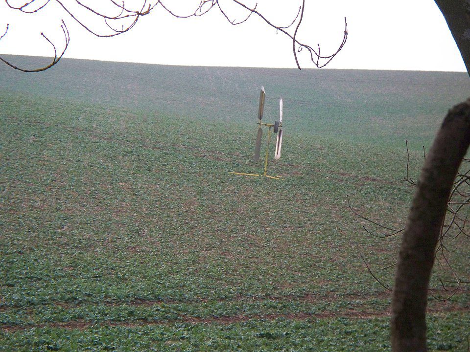 Rotating rustic bird scarer in the middle of a crop field, simple metal and wood structure, designed to spin and create noise, placed in open farmland to deter birds, surrounded by green low vegetation, slightly foggy background