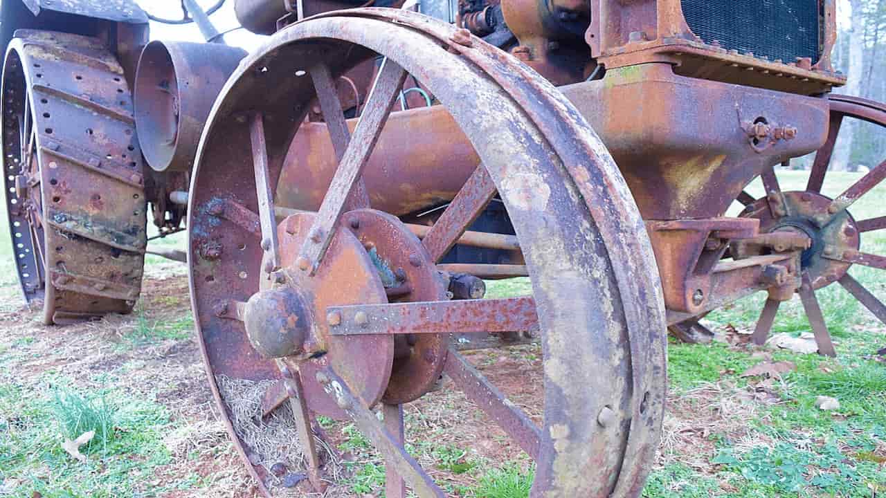 Close-up of a rusted, antique tractor wheel, showing corroded metal, bolts, and spokes, set on patchy grass
