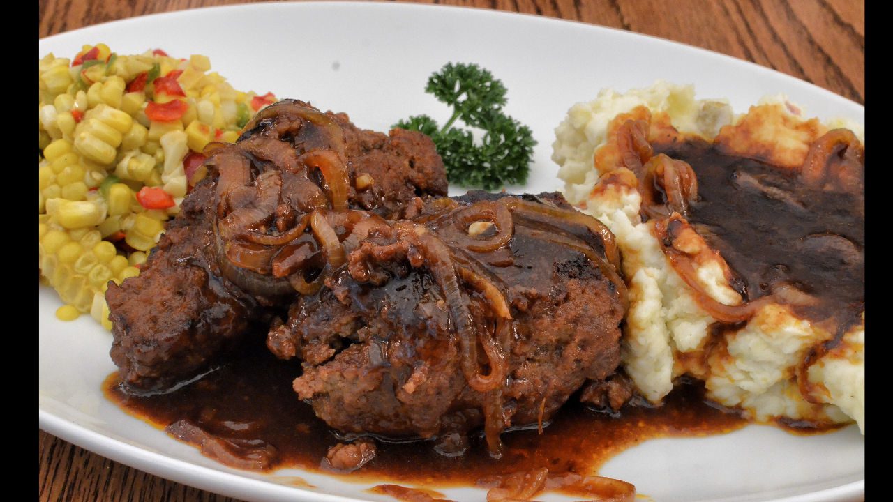 Salisbury steak with caramelized onions and brown gravy, served with mashed potatoes and seasoned corn medley on a white plate